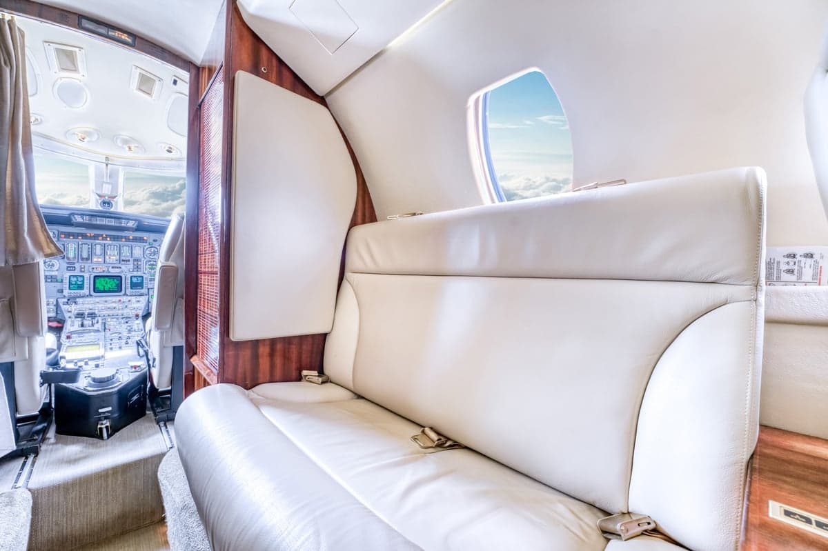 The aircraft interior shows a white leather bench seat and a view into the cockpit.