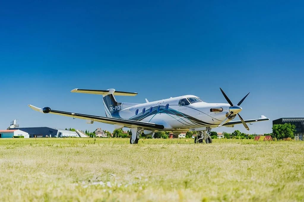 A white Pilatus PC-12 aircraft with blue and black accents is parked on a grassy field with buildings in the background.