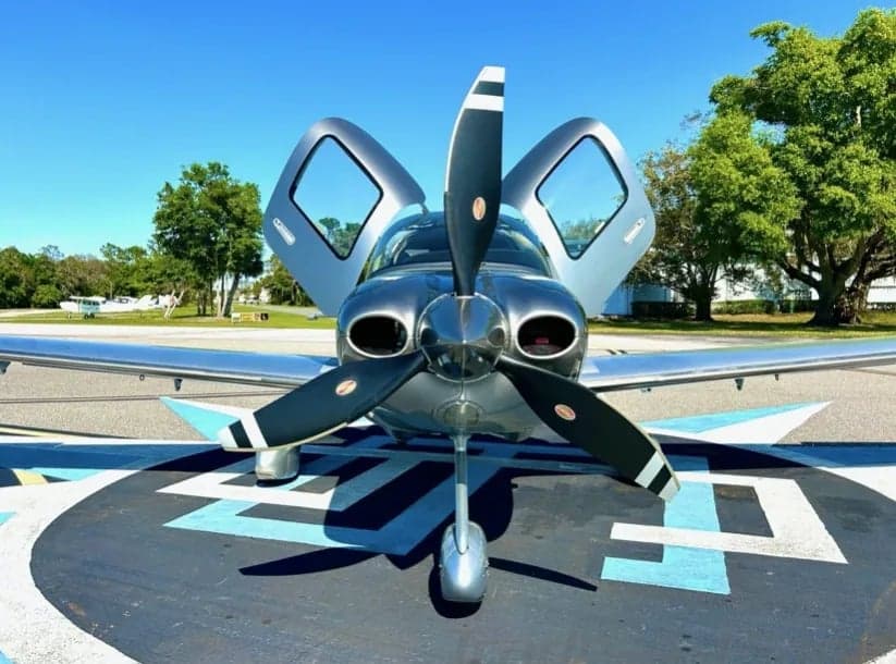 Front view of the silver Cirrus aircraft with its doors open and propeller visible.