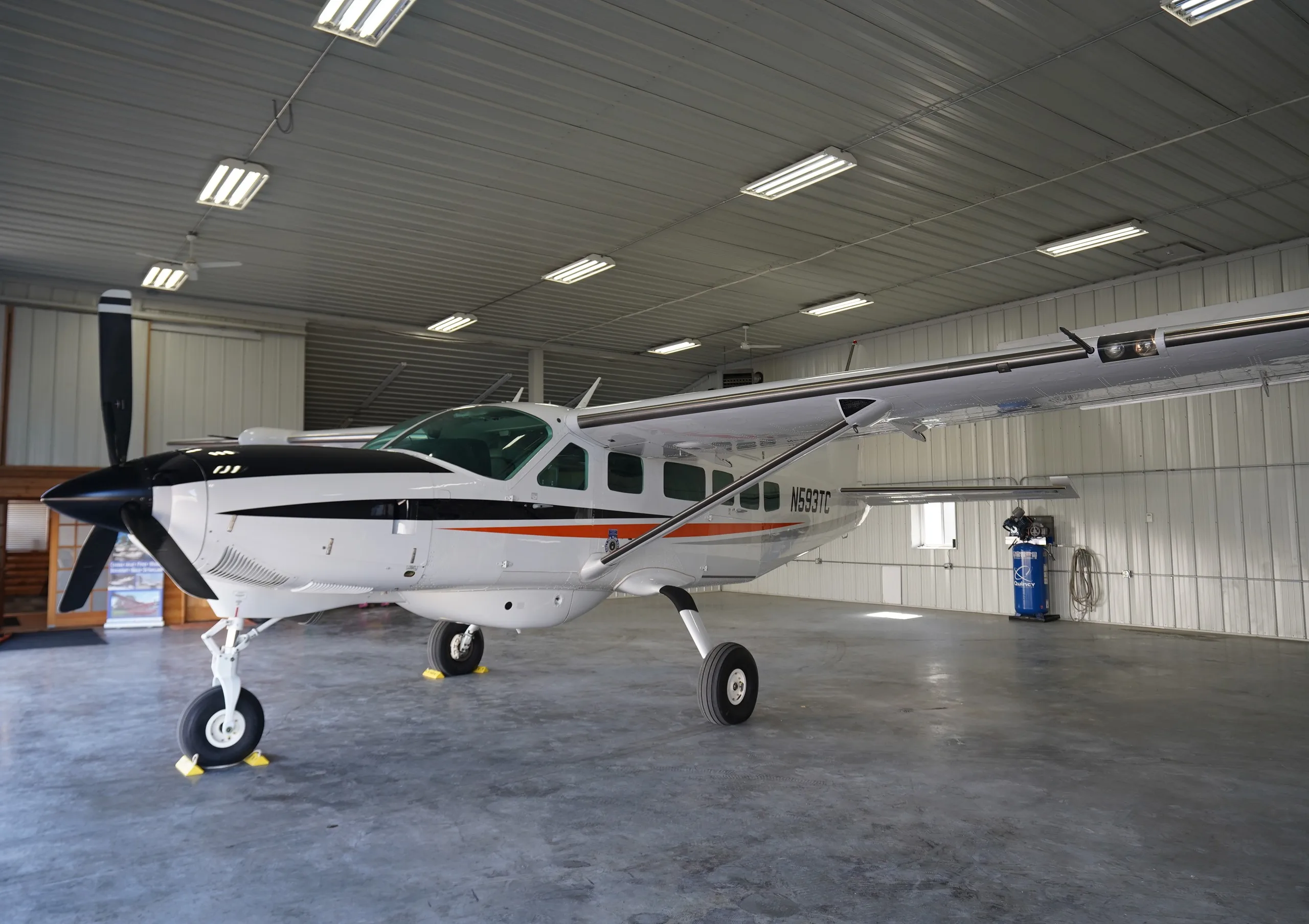 A white Cessna Caravan (N593TC) with black and orange stripes parked inside a well-lit hangar.