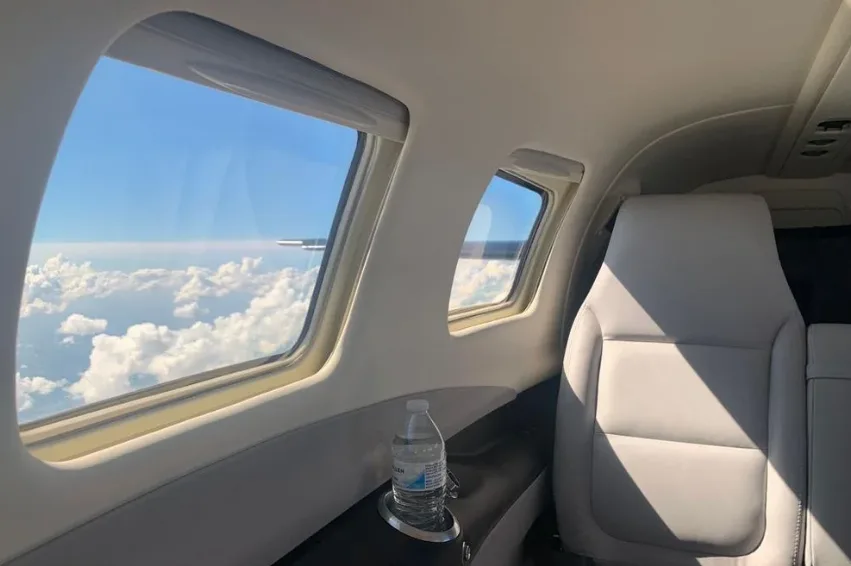 The aircraft's cabin interior with a light grey seat and windows offering a view of clouds.