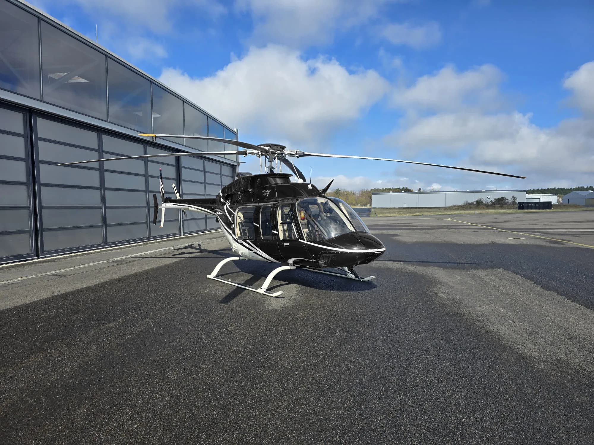 A black and white helicopter, possibly a Bell 505, parked on an asphalt apron next to a modern hangar under a partly cloudy sky.