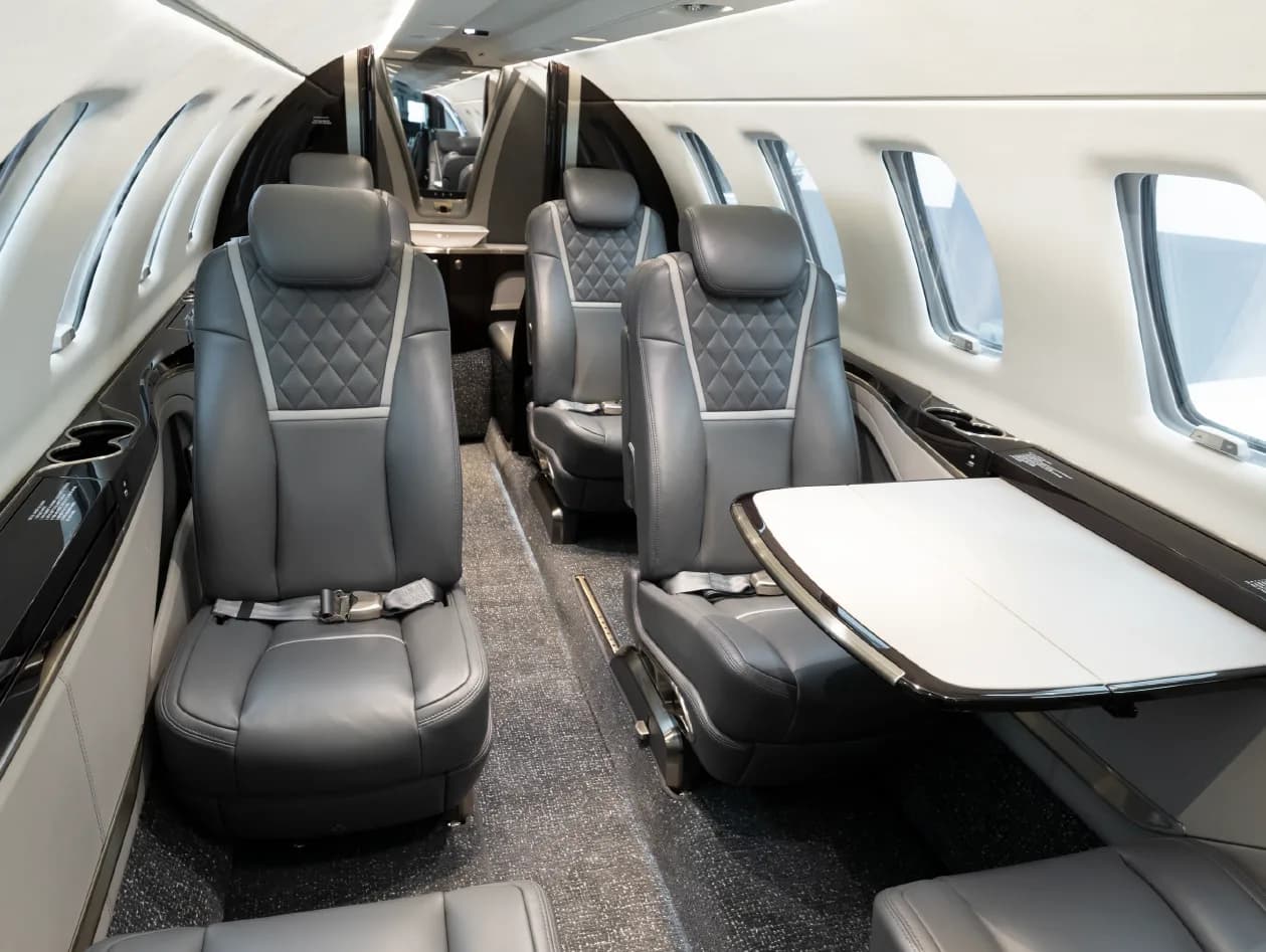 The luxurious interior cabin of a business jet, featuring grey leather seats with diamond stitching and pull-out tables.