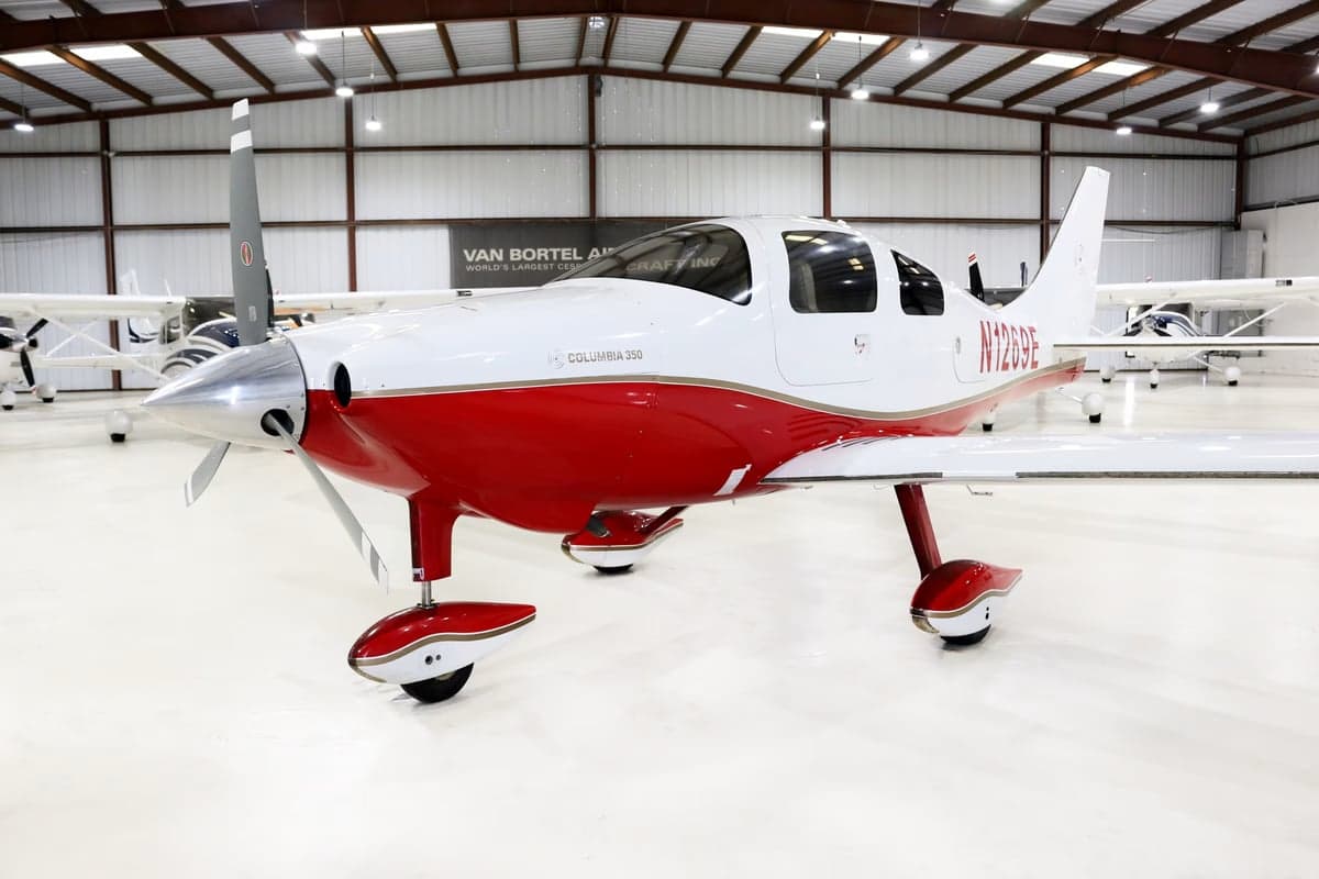 A Columbia 350 aircraft with red and white paint scheme is parked in a hangar.
