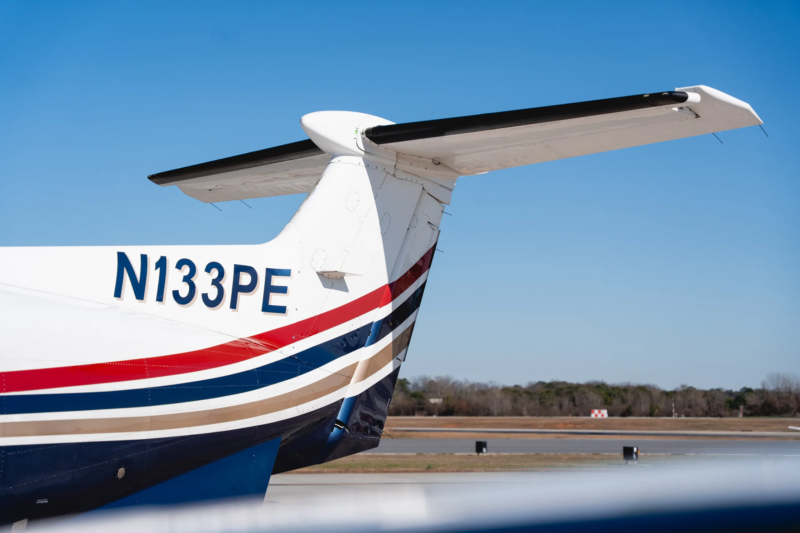 A close-up view of the tail section of the Pilatus PC-12 aircraft, registration N133PE, showing the blue, red, and gold stripe paint scheme.