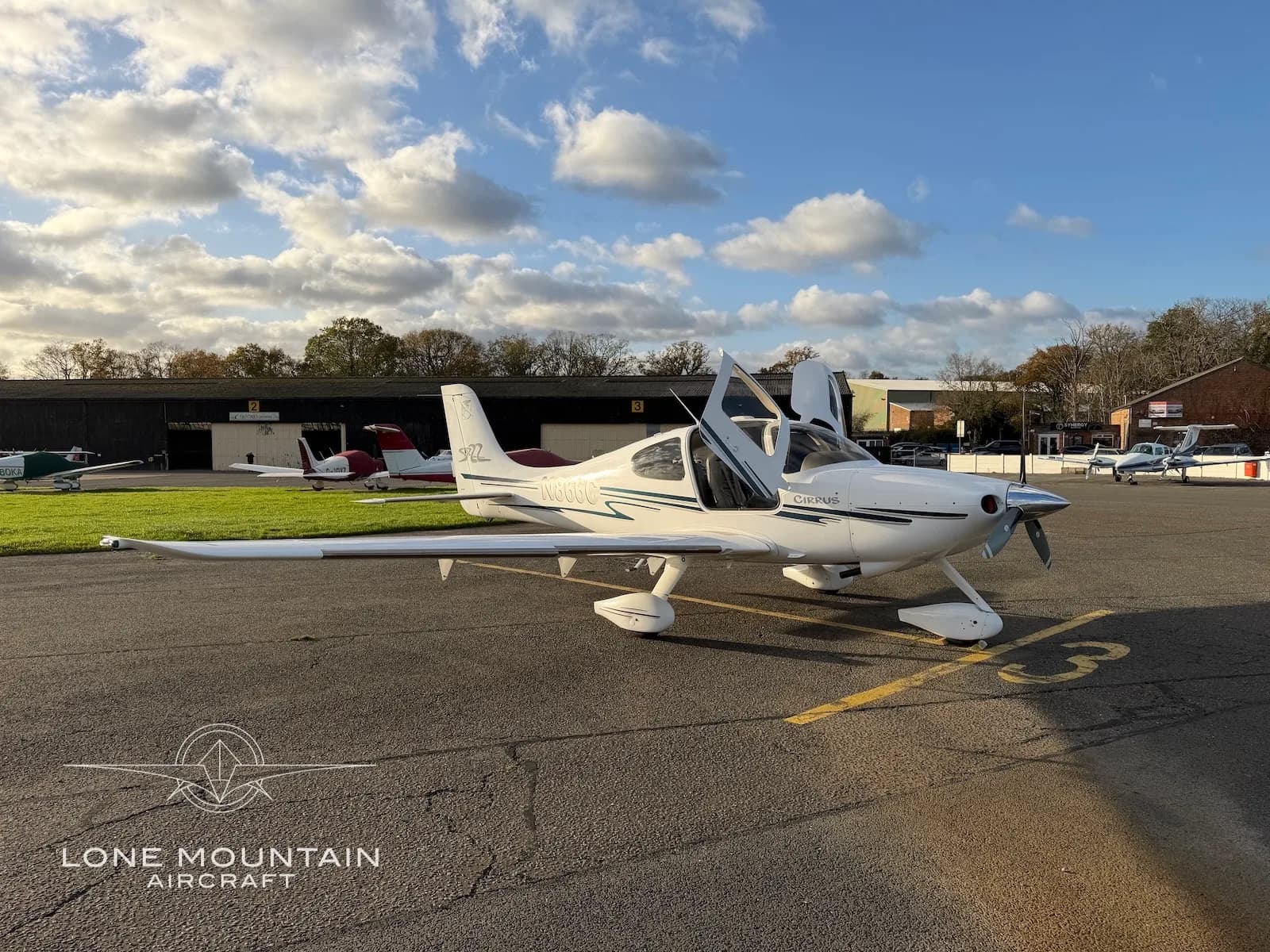 A three-quarter front view of a white Cirrus SR22 aircraft with black and grey stripes, registration N866C, parked on an asphalt apron with both doors open.