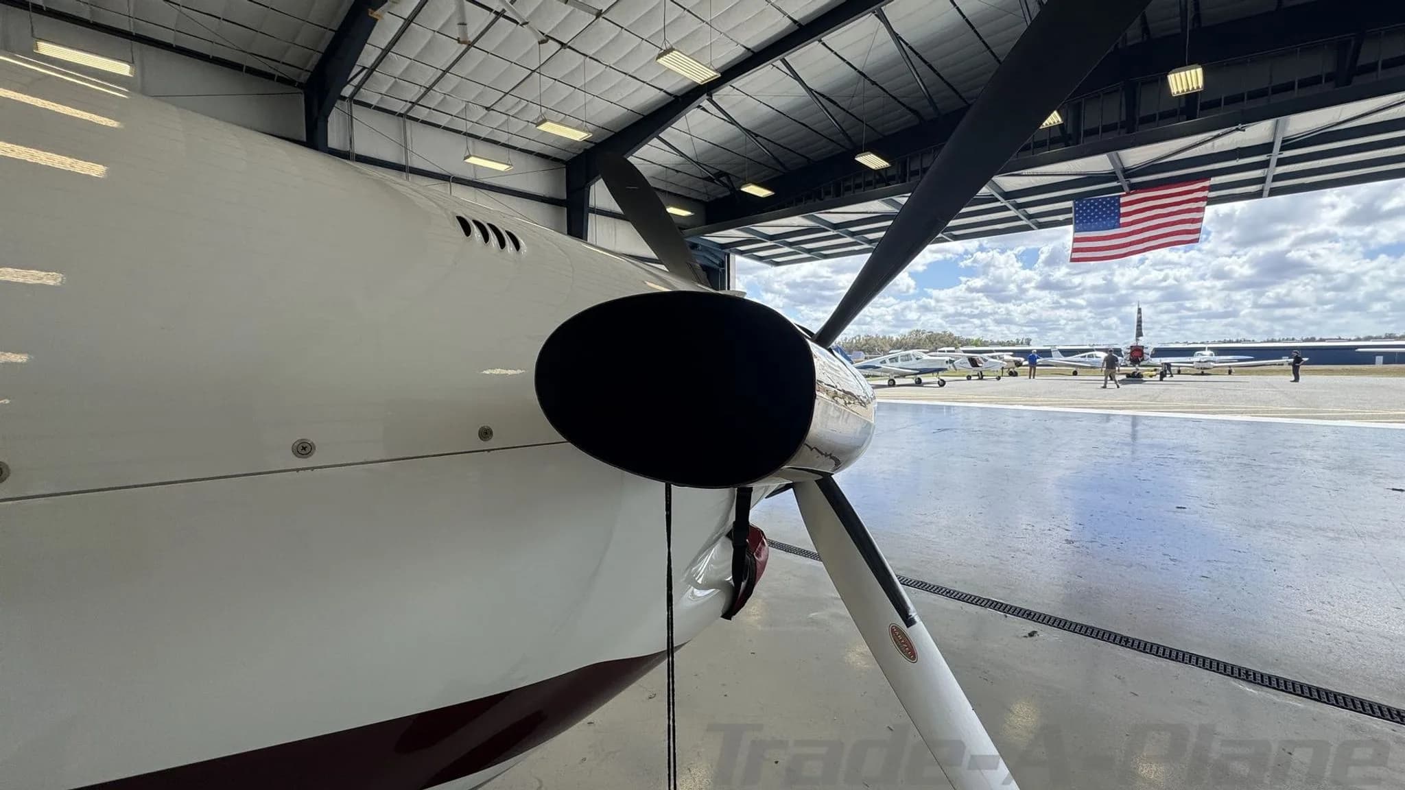 Close-up of the propeller and nose of a white Epic LT aircraft, with other aircraft and an American flag visible outside a hangar.
