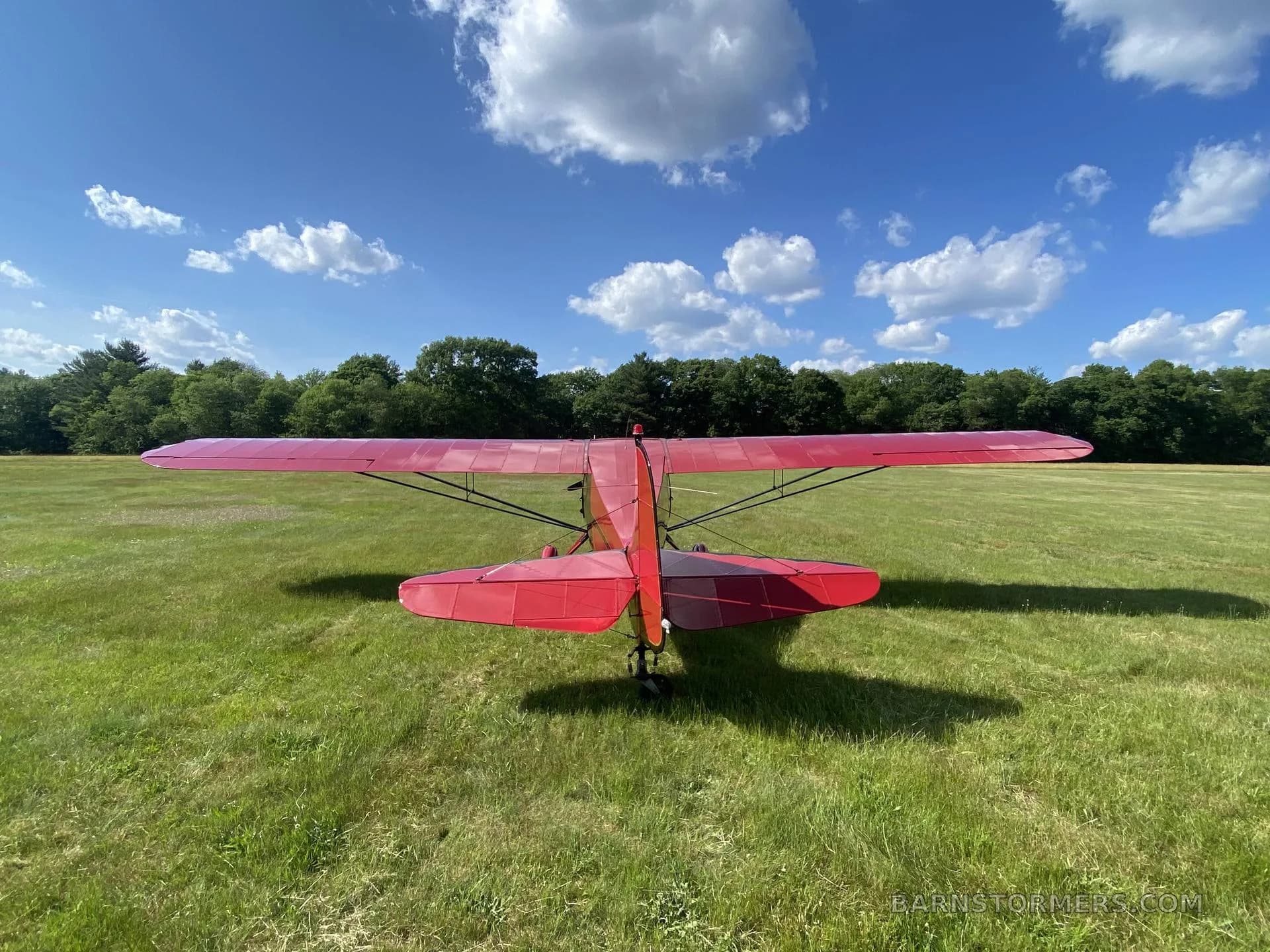 The rear view of a red single-engine high-wing aircraft parked on a grassy field under a blue sky.