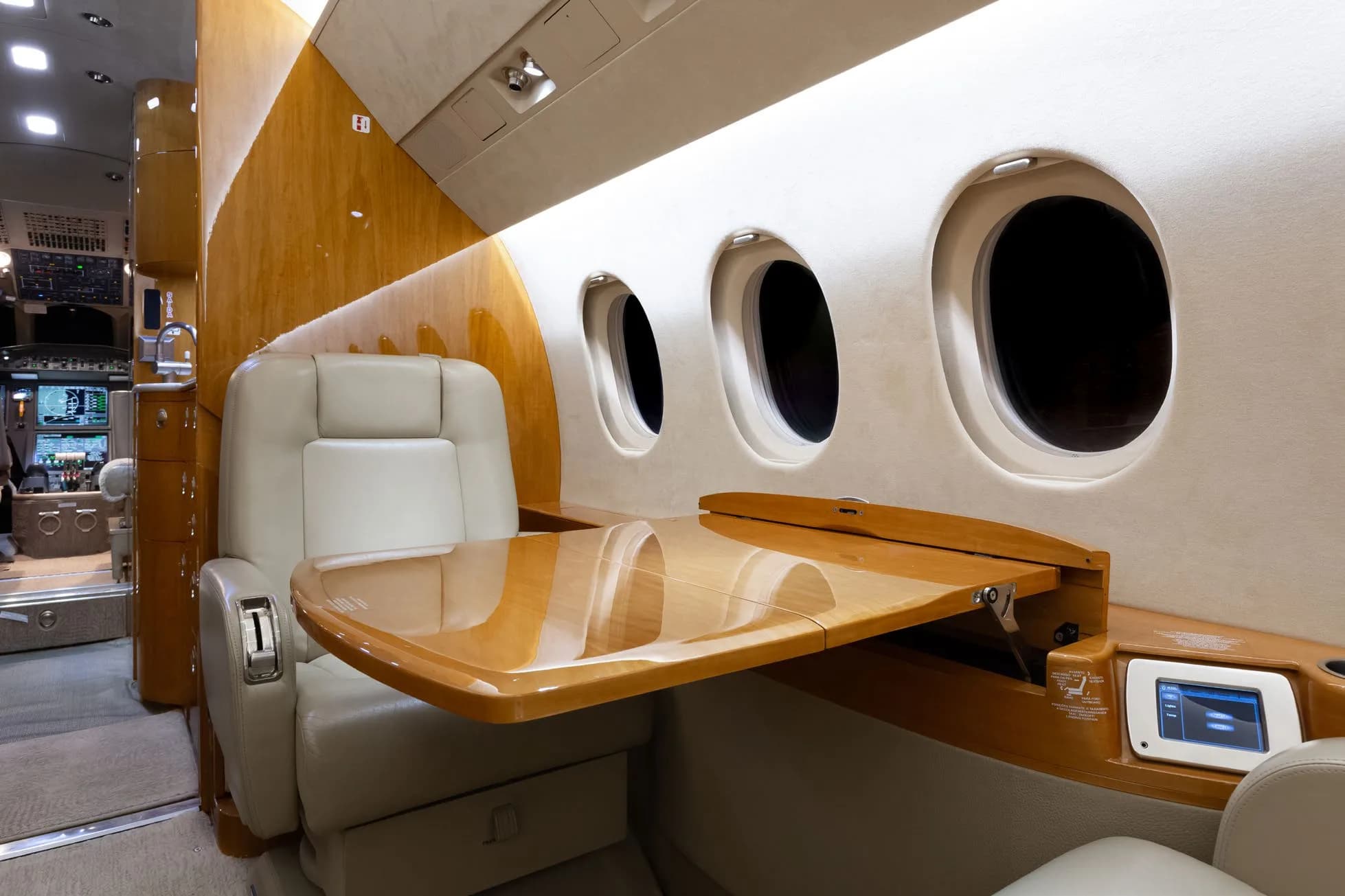 A close-up view of a beige leather seat and a polished wooden fold-out table next to windows in a private jet cabin.
