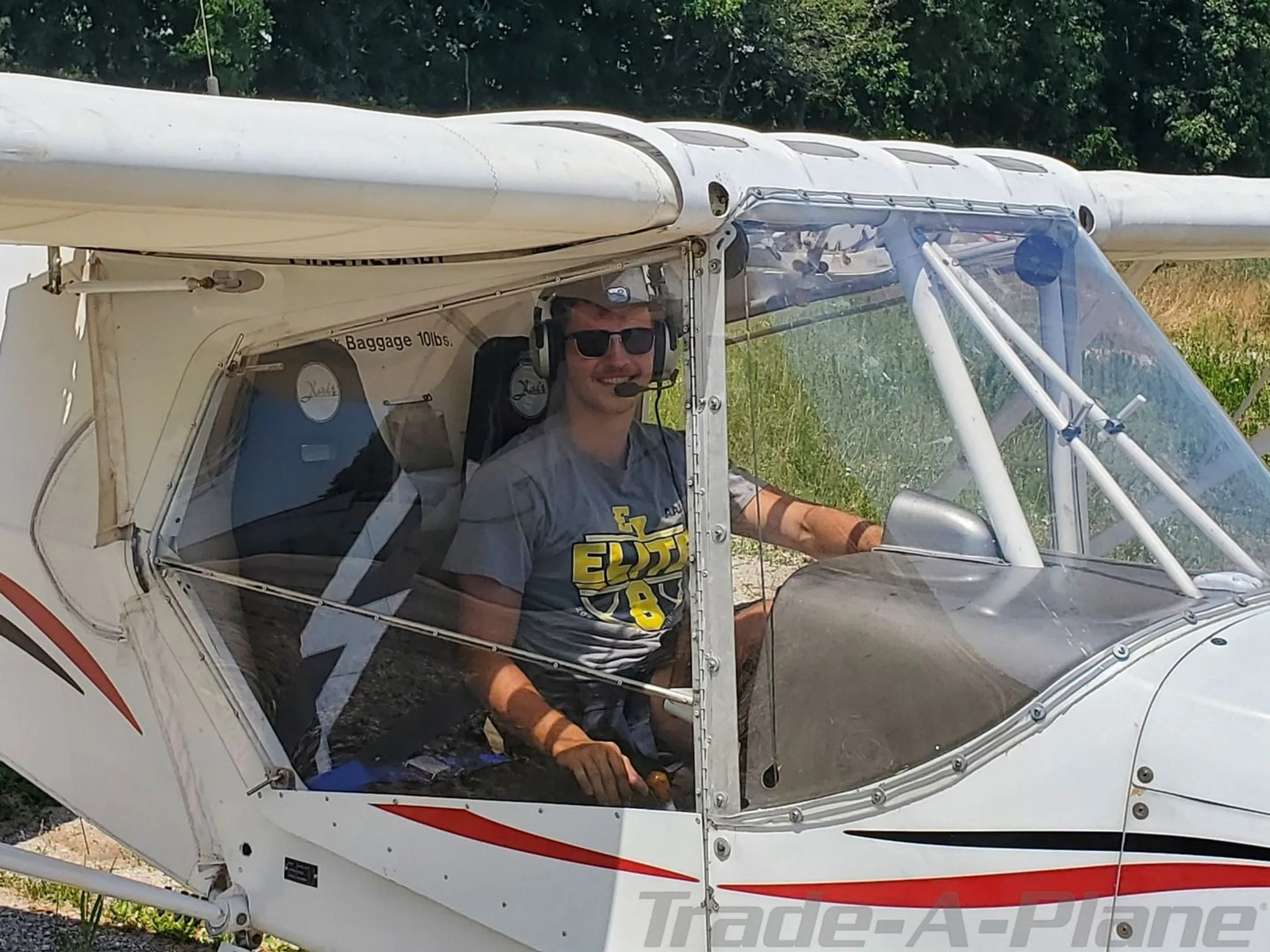 A smiling pilot wearing sunglasses and a headset sits in the cockpit of the N17XA X-Air aircraft.