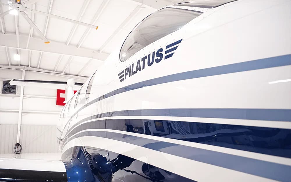 A close-up view of the white and blue fuselage of a Pilatus PC-12 aircraft, showing the "PILATUS" logo.