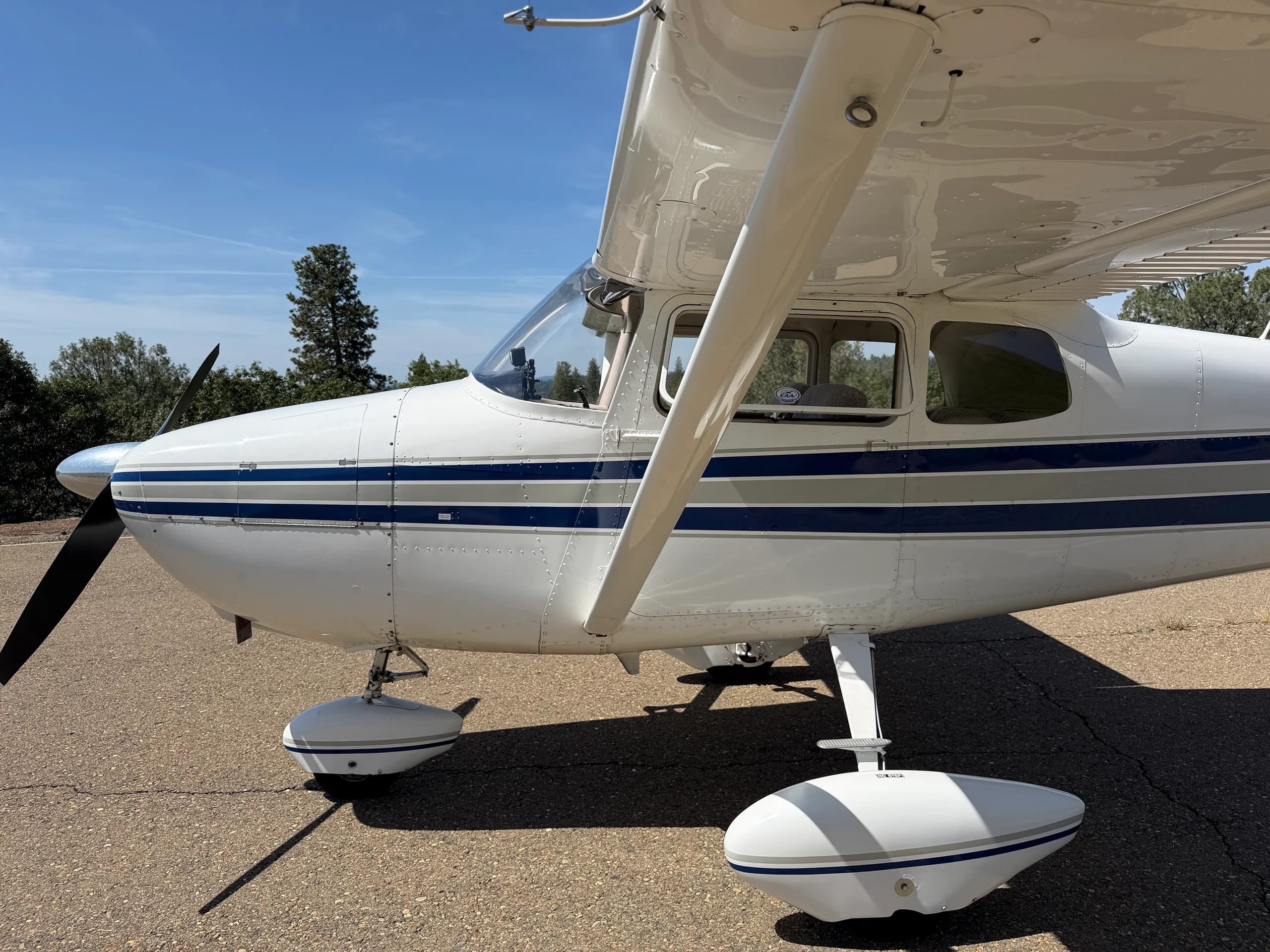 A close-up side view of the front fuselage of a white Cessna 172, showing the propeller, landing gear, and blue stripe.