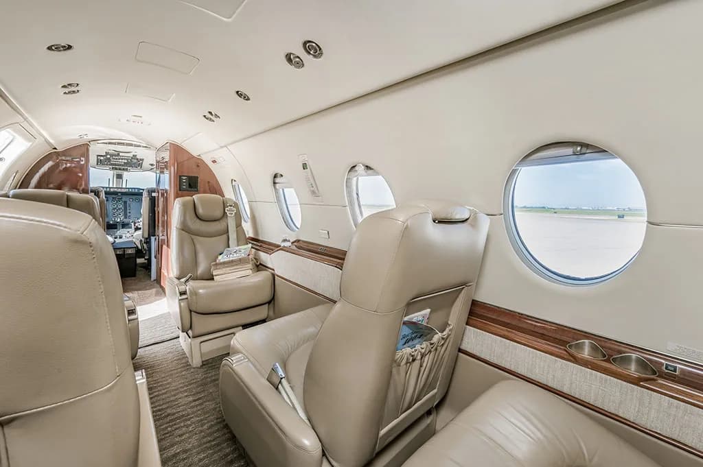 The interior cabin of a business jet with beige leather seats and large windows, looking forward towards the cockpit.