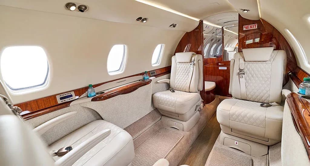 A side view of the private jet cabin interior, highlighting the light-colored seats, polished wood trim, and a lavatory entrance.