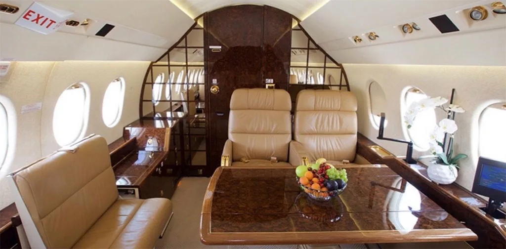 The rear section of a private jet cabin, showing tan leather seats, a polished wood table with fruit, and an "EXIT" sign.