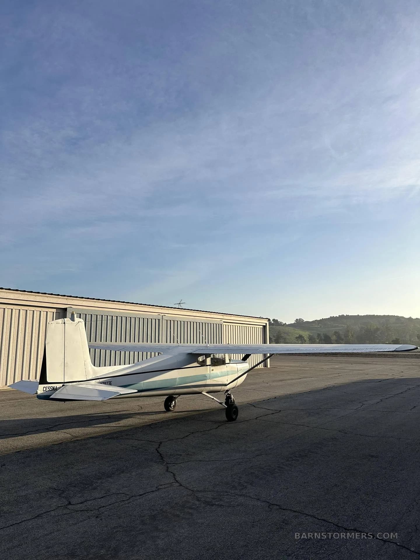A white and blue Cessna 150/152 aircraft is parked on a tarmac next to hangars with green hills in the distance.