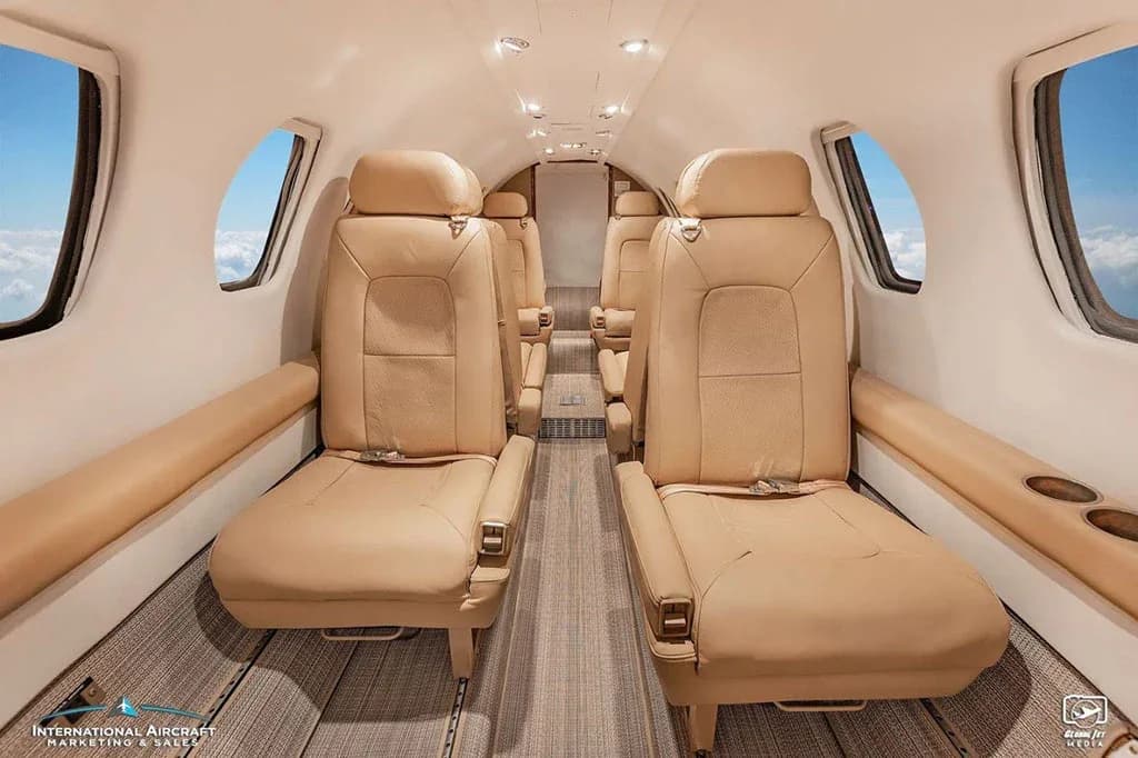 A view down the aisle of a business jet cabin with beige leather seats and windows showing clouds.