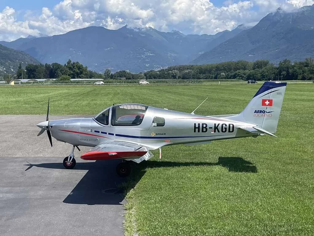 A silver Sonaca 200 aircraft (HB-KGD) with red wingtips is parked on a grassy airfield with mountains in the distance.