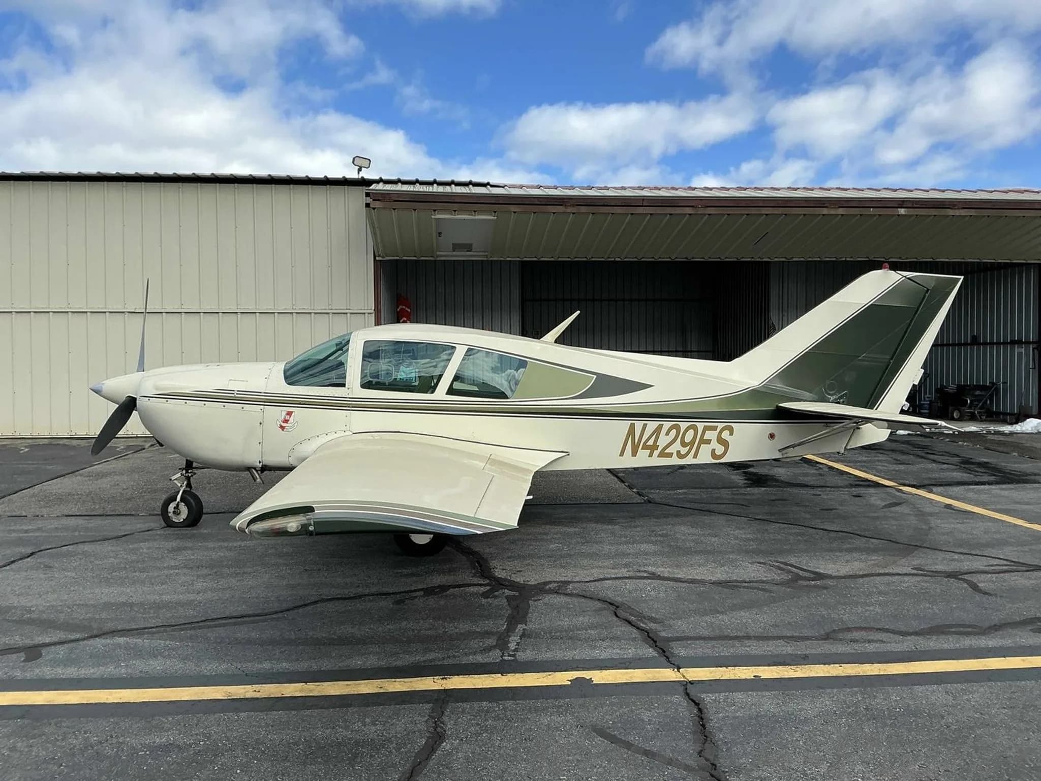 A side view of the white and green Bellanca Viking aircraft, N429FS, parked in front of an open hangar.