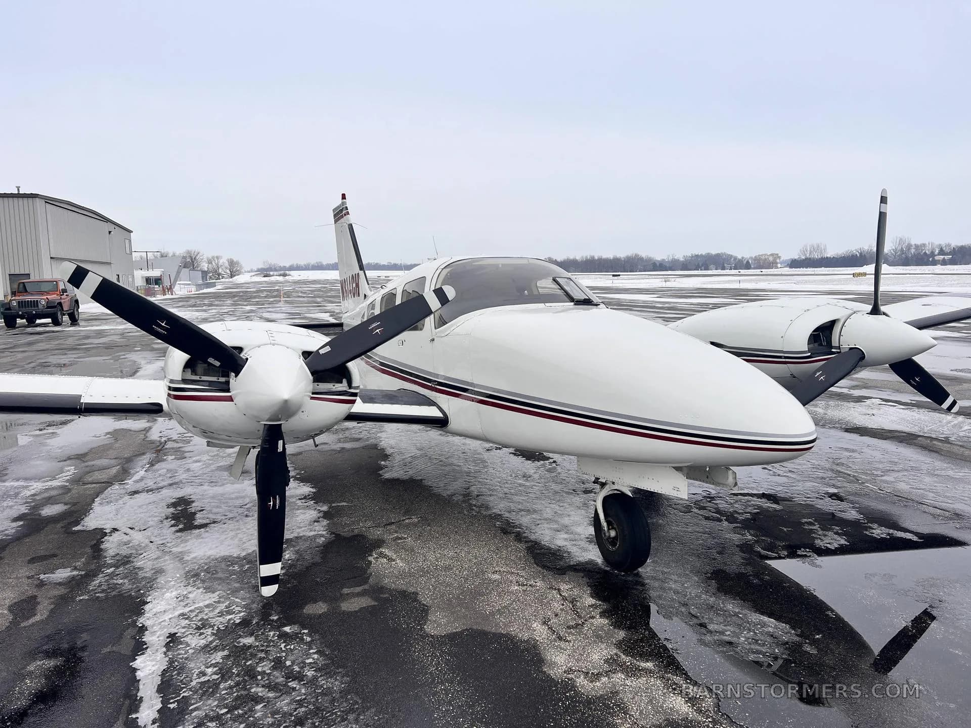 A front-quarter view of a white twin-engine Piper aircraft with maroon and black stripes, tail number N814CH, parked on a wet tarmac.