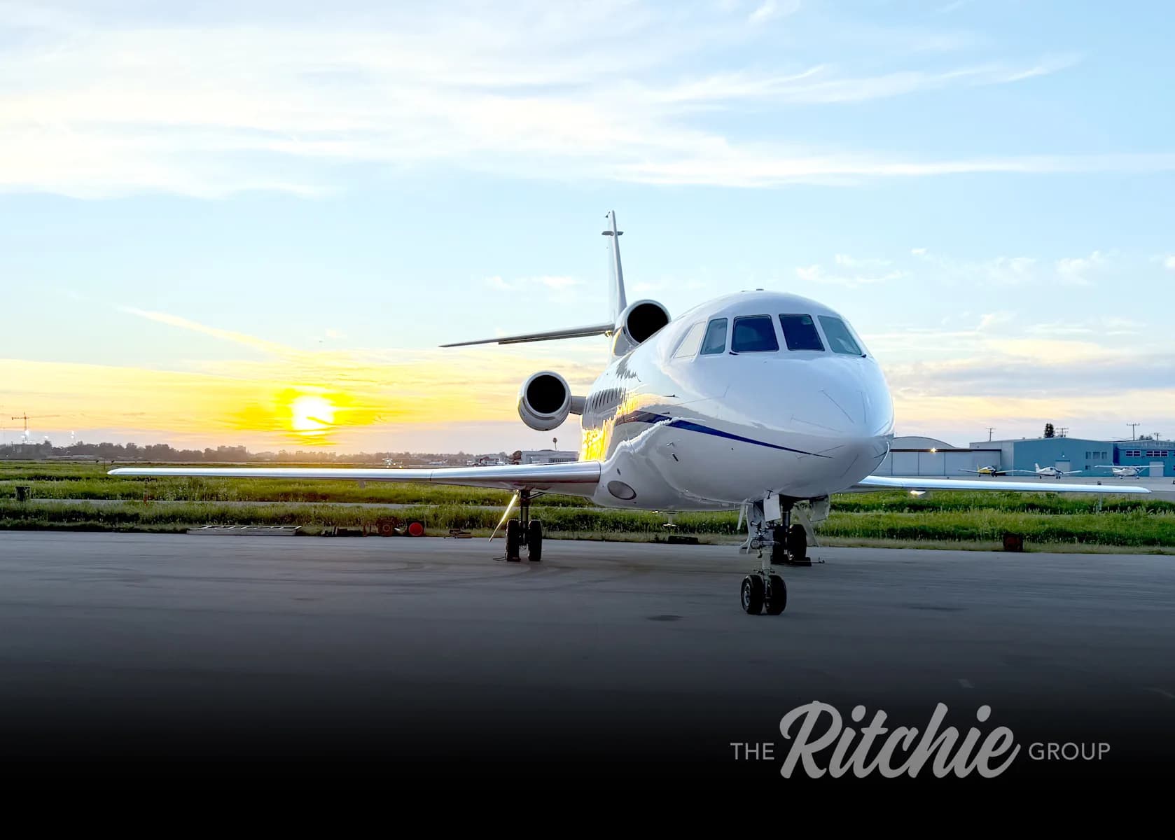 A white business jet with blue accents, viewed from the front, parked on an airfield at sunrise.