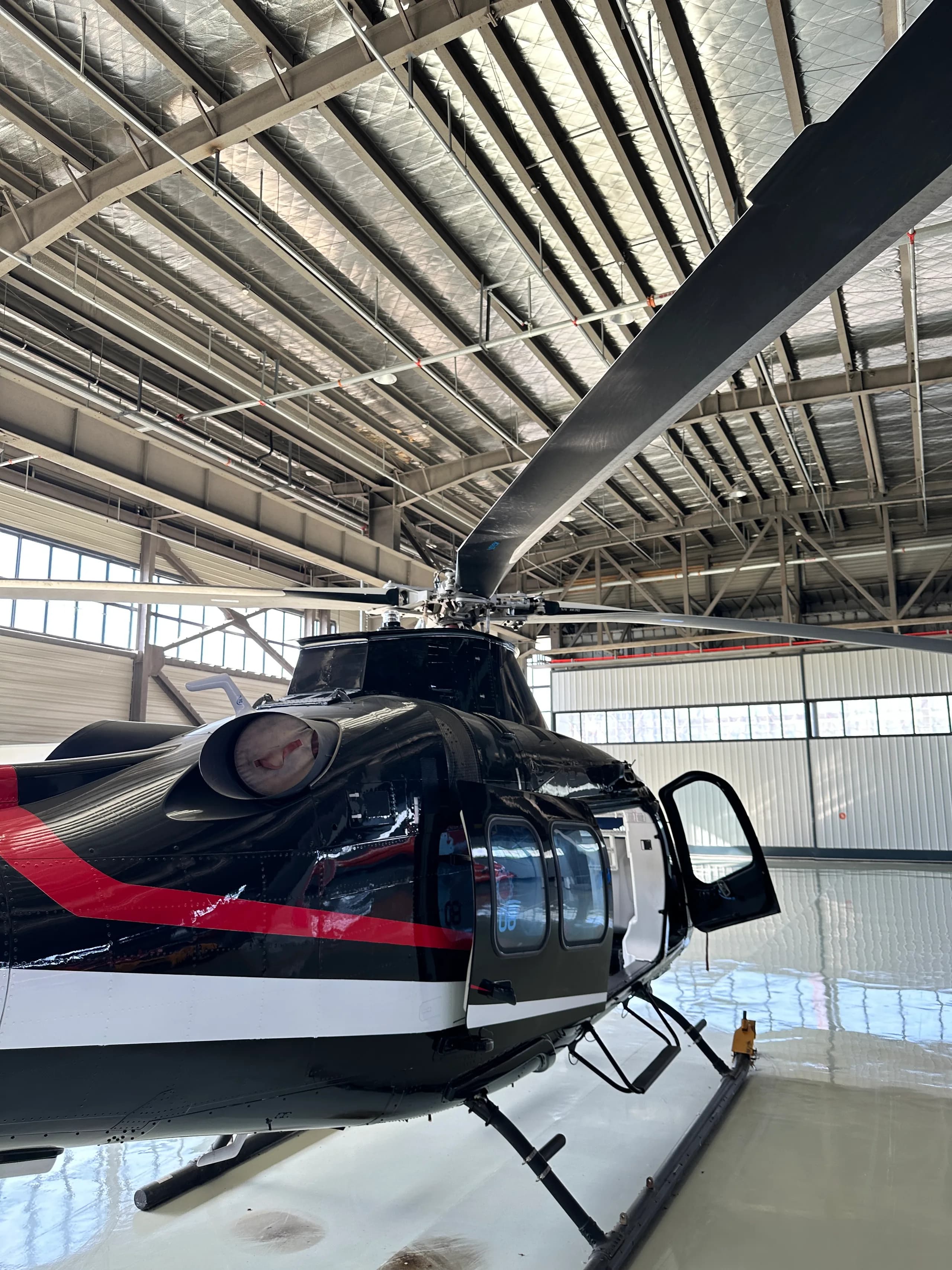 The rear-left side of a black helicopter with red and white stripes is shown in a hangar, with the passenger door open.