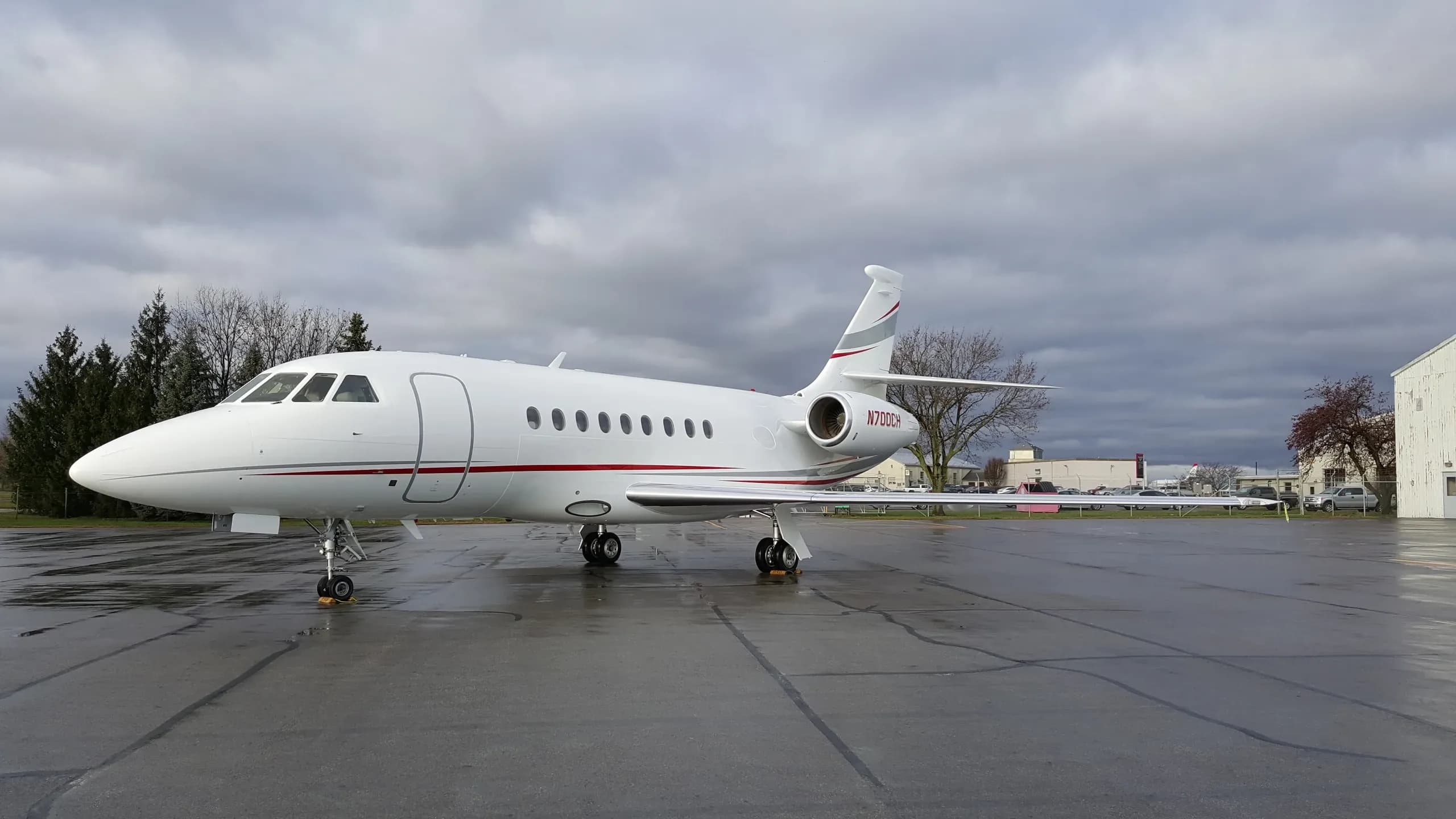 A white Dassault Falcon 2000 business jet, registration N700CH, with red and grey stripes, parked on a wet tarmac.