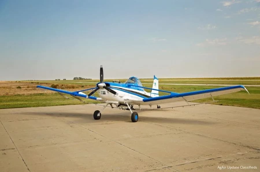 A white and blue agricultural aircraft with a large propeller parked on an airfield runway under a clear sky.