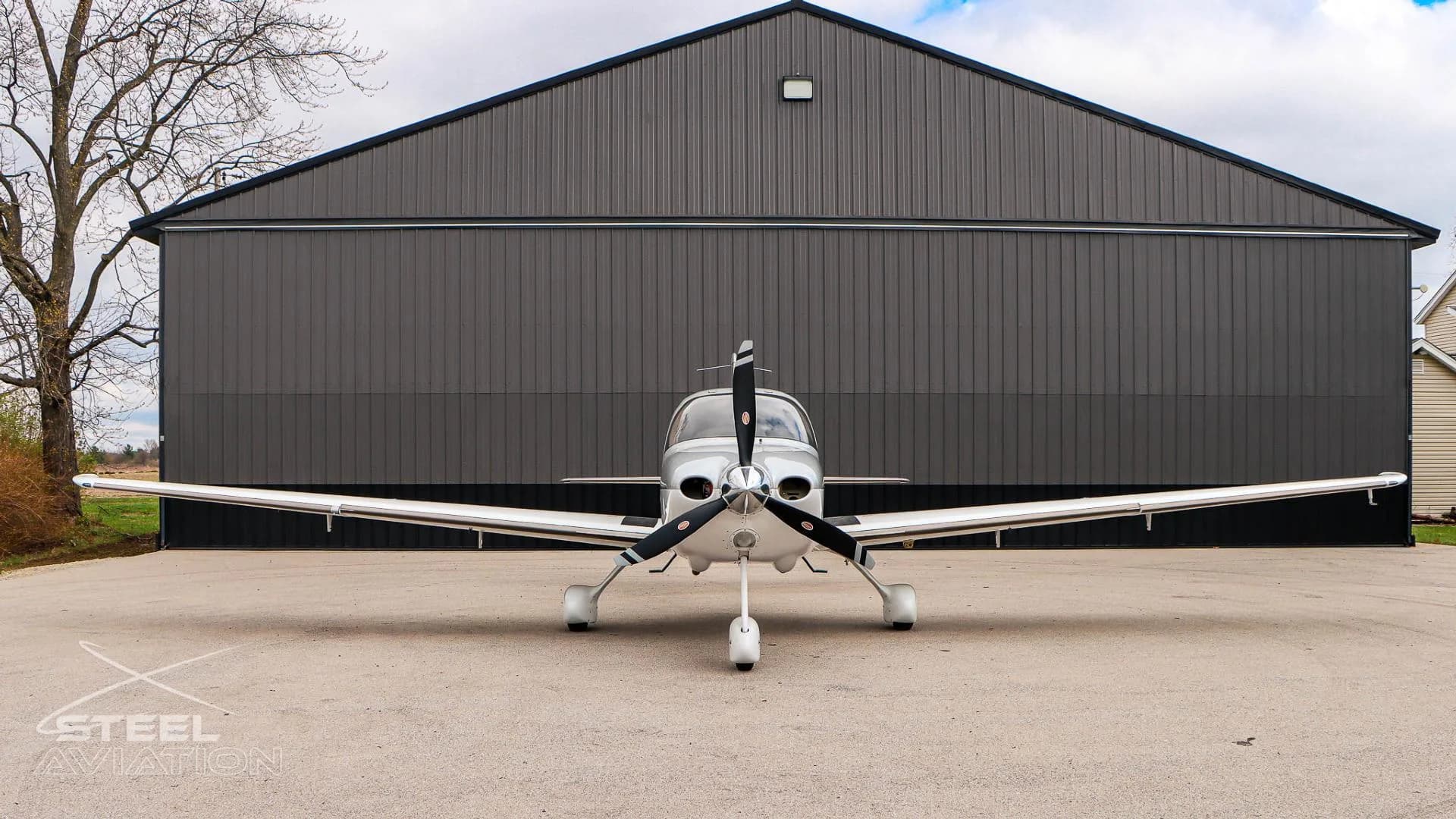 A front view of a white and silver Cirrus SR22 aircraft parked on an asphalt apron in front of a dark grey hangar.