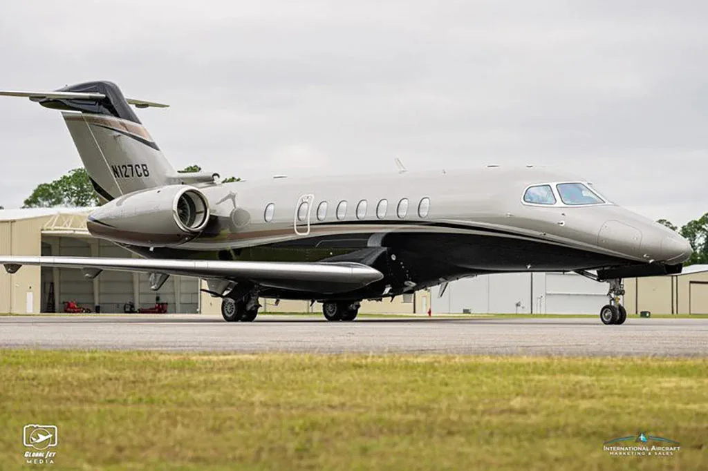 A side view of a grey and black business jet with registration N127CB on a tarmac next to a grassy area.