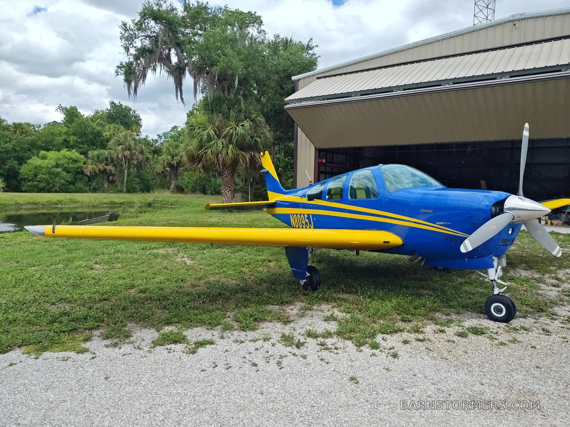 A blue and yellow single-engine aircraft is parked on a gravel path next to a hangar, with trees and a pond in the background.