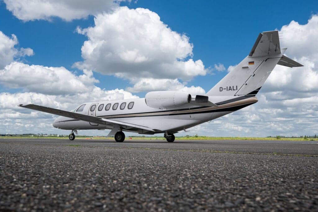 A white Cessna Citation CJ3 (D-IALI) private jet with black and gold stripes, showing the tail and engines, parked on an asphalt runway under a blue sky with white clouds.