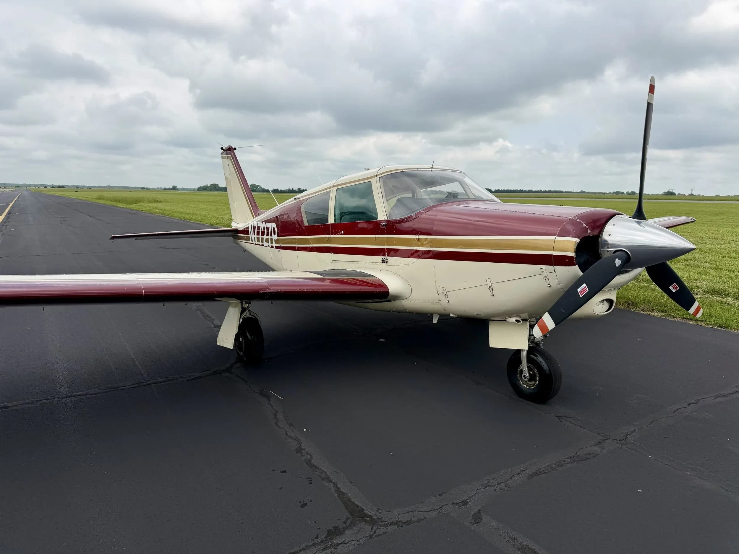 Front-side view of a maroon and white Piper Comanche N7727P parked on an asphalt runway.