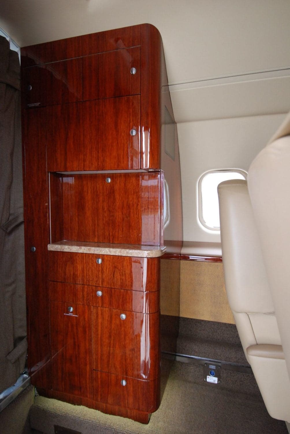 A polished dark wood cabinet with a light countertop inside a business jet's cabin, next to a window.