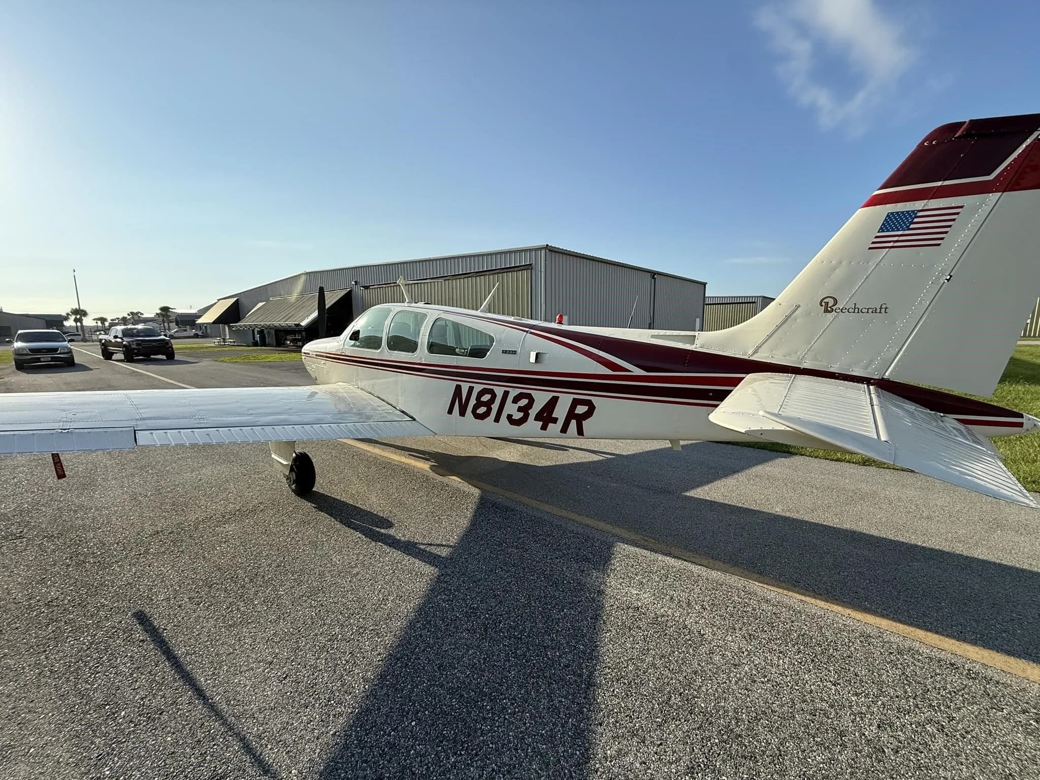 A white Beechcraft aircraft with maroon stripes, tail number N8134R, is parked on an asphalt ramp with hangars in the background.