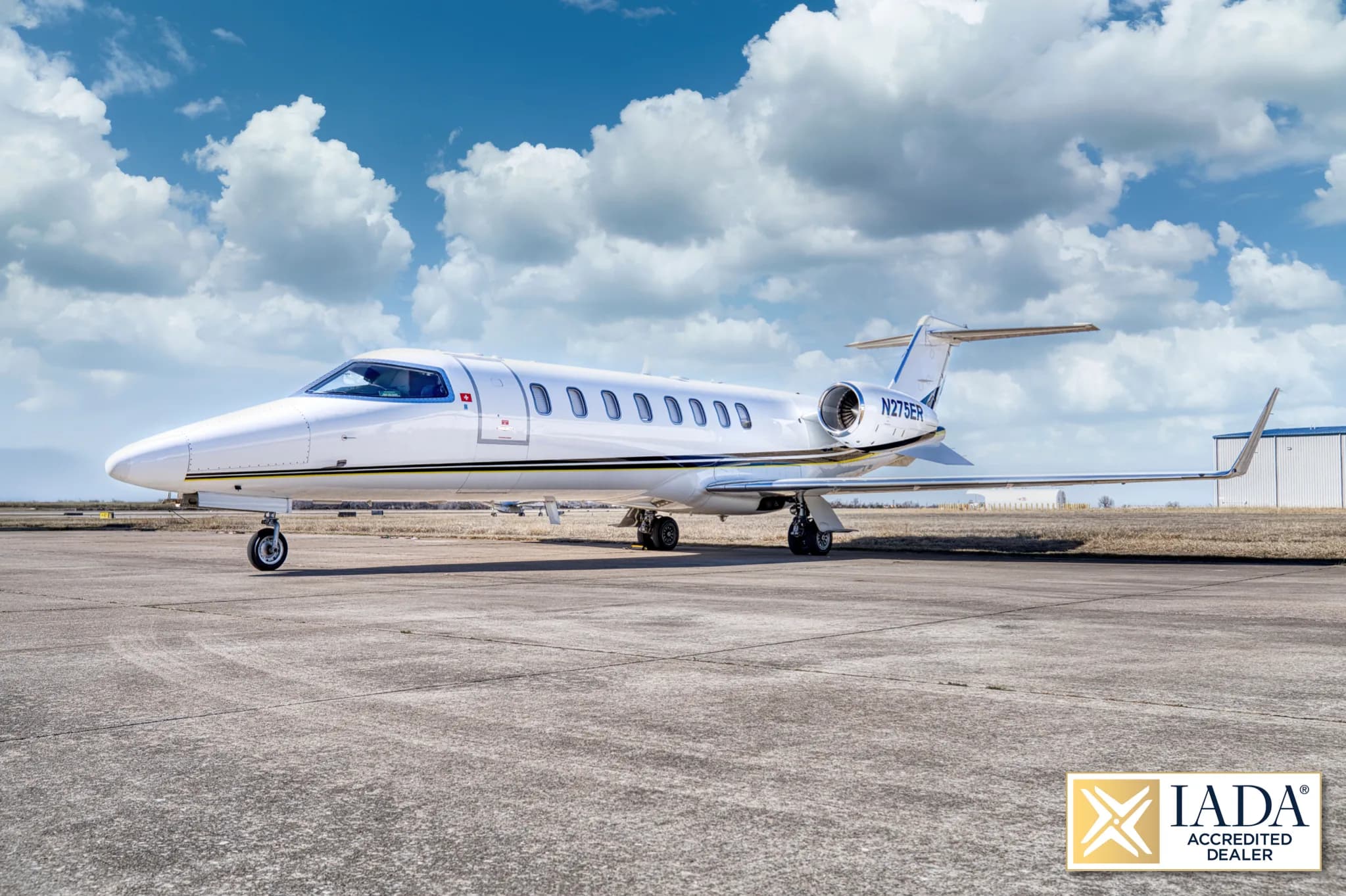 A white Learjet 60 (N275ER) with a black and yellow stripe, parked on a tarmac with a hangar in the background.