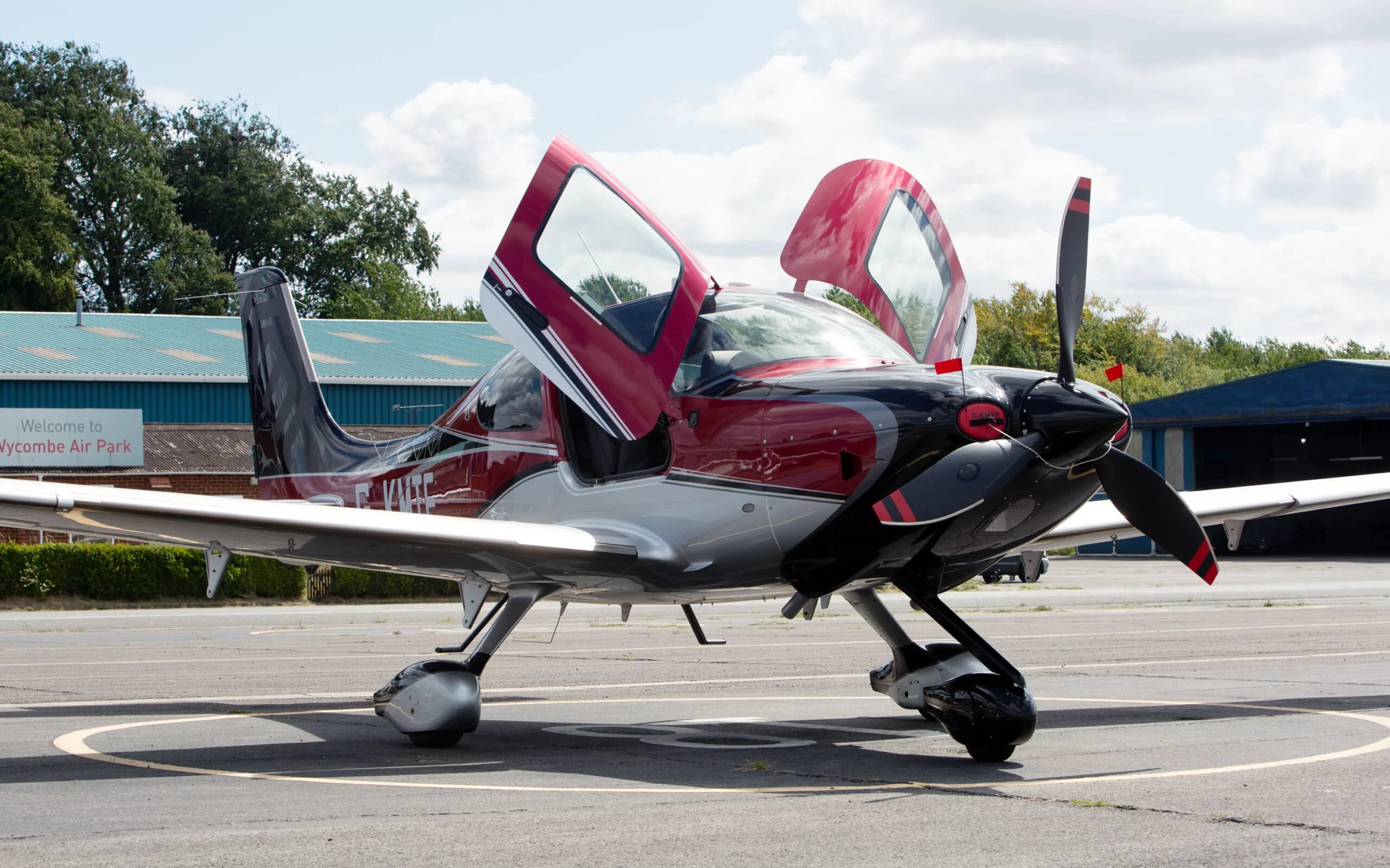 A red, black, and silver Cirrus SR22 aircraft with its gull-wing doors open, parked on an airfield with a 'Welcome to Wycombe Air Park' sign in the background.