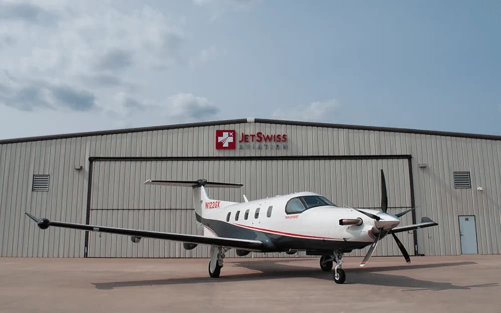 A white and grey Pilatus PC-12 turboprop aircraft with tail number N122GX parked on the tarmac in front of a hangar under a cloudy sky.