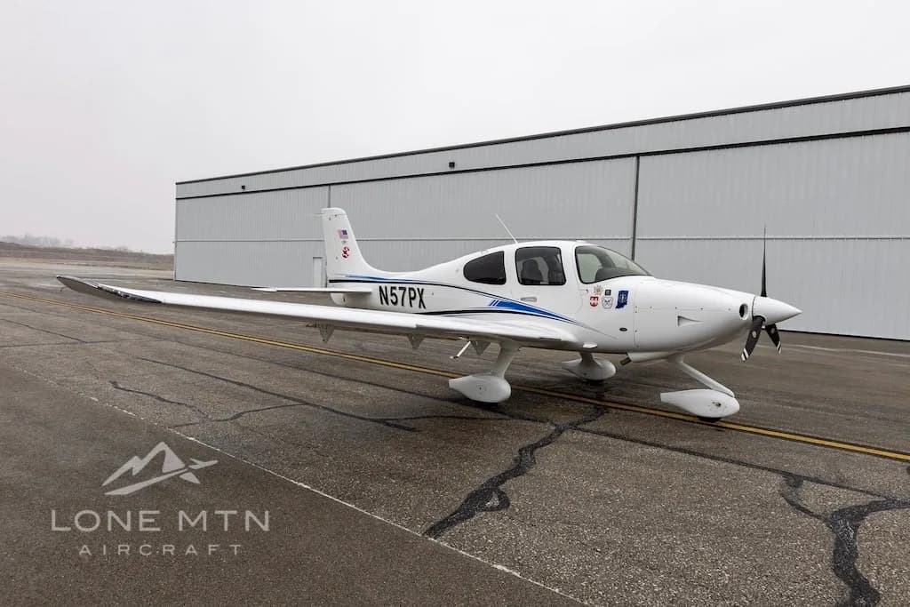 A white Cirrus SR22 aircraft with blue and black stripes, tail number N57PX, parked on an airport tarmac in front of a hangar.