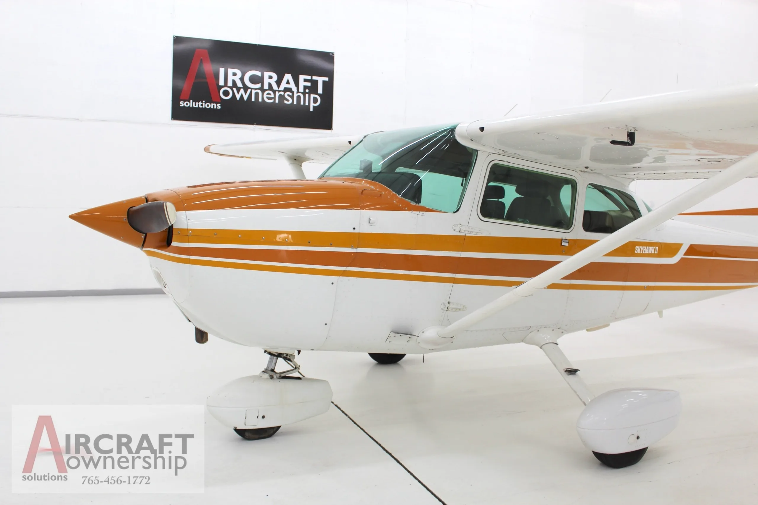 A white and orange Cessna Skyhawk II (N5546J) is parked in a brightly lit hangar, viewed from the front-side.