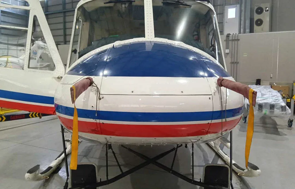 A direct front view of a white helicopter with a blue nose and red and blue stripes, parked in a hangar.