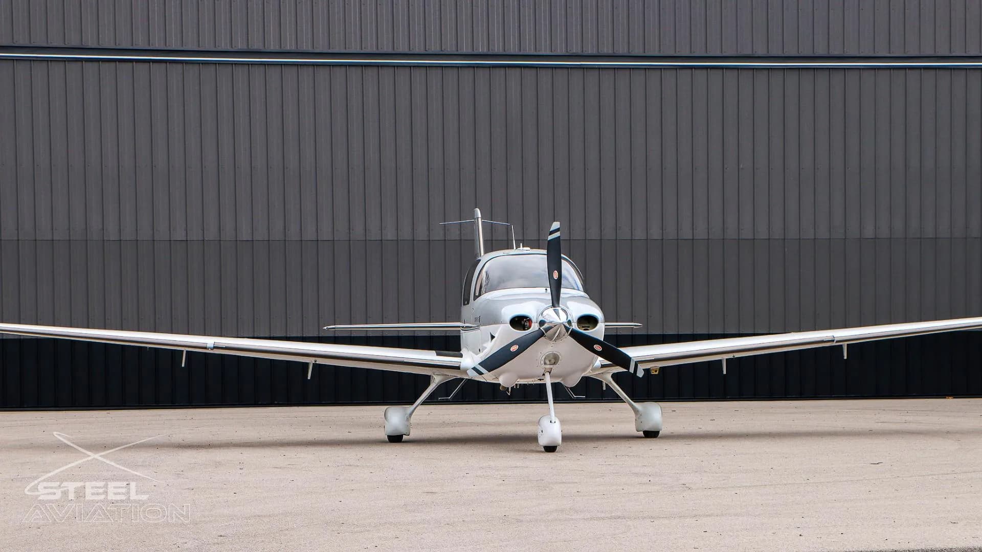 A front view of a white and silver Cirrus SR22 aircraft parked on an asphalt apron in front of a dark grey hangar.