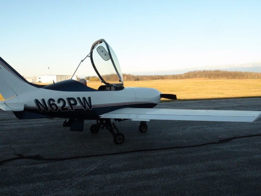 Side view of a white and blue aircraft with the canopy open on a paved surface.