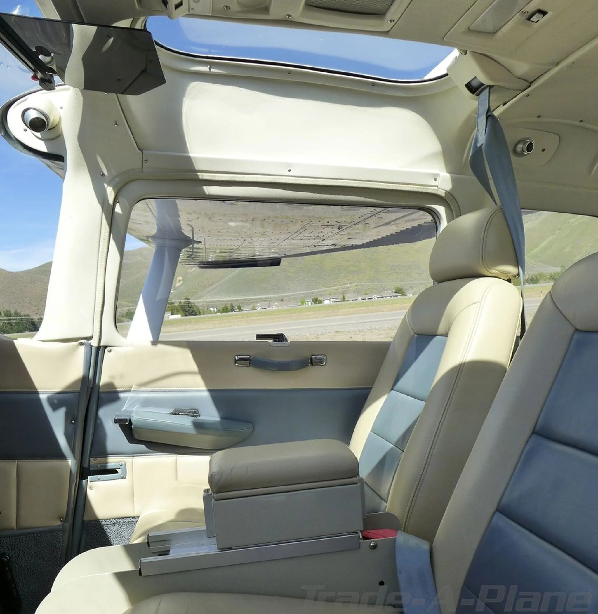 The interior cabin of a Cessna 182 aircraft, showing beige and blue seats and a view out the window to a runway and hills.
