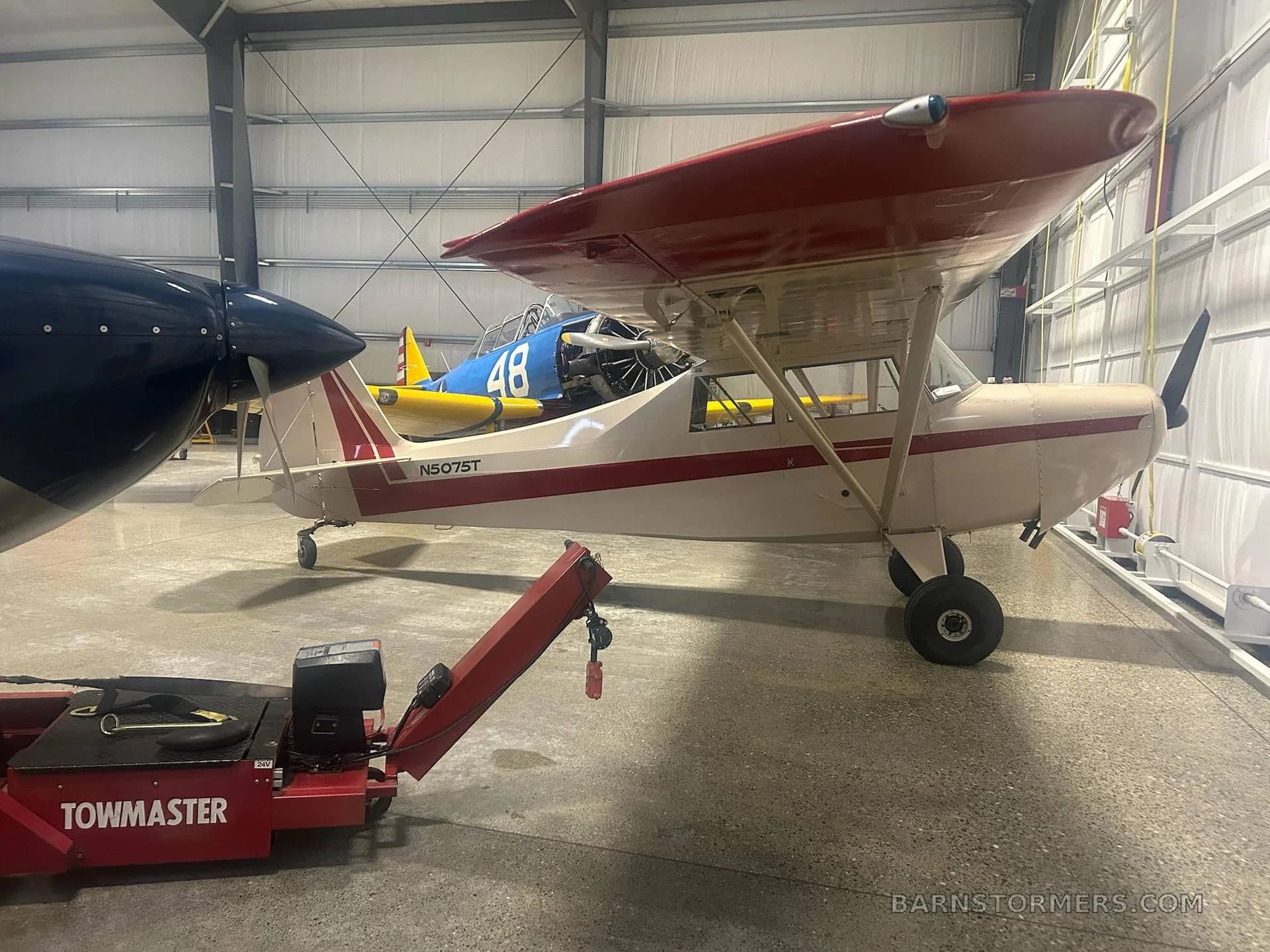 A white and red single-engine taildragger aircraft (N5075T) parked inside a hangar next to a blue aircraft and a red towmaster.