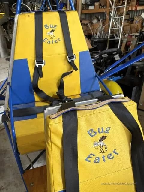 The yellow and blue fabric seat of an ultralight aircraft with "Bug Eater" embroidered on it, showing seatbelts.