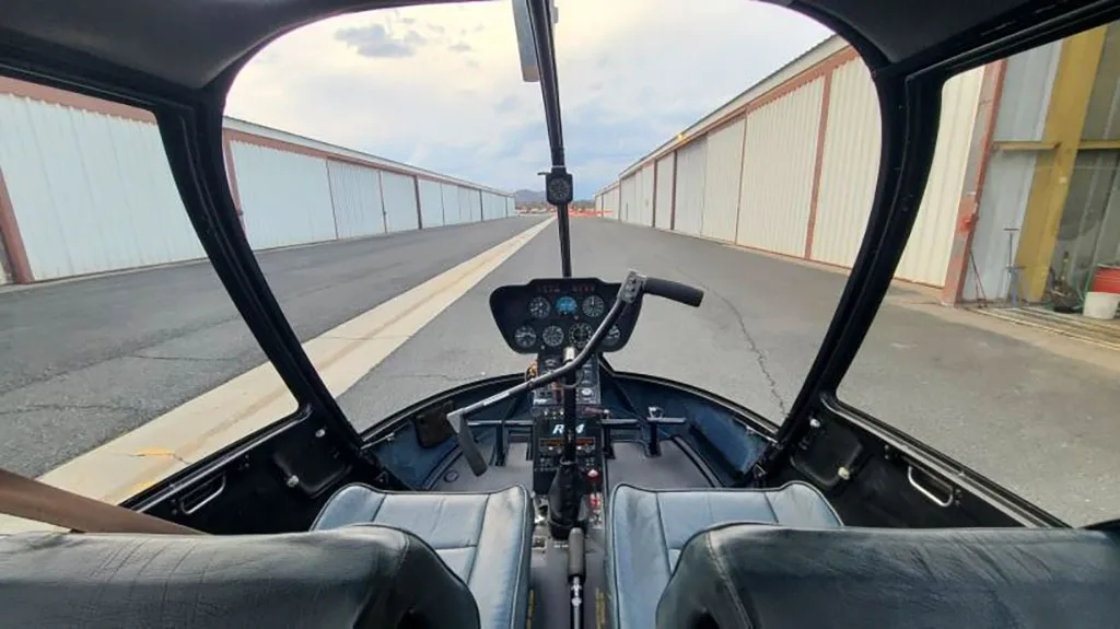 View from inside the cockpit of a Robinson R44 helicopter, showing the instrument panel, seats, and an airfield runway ahead.