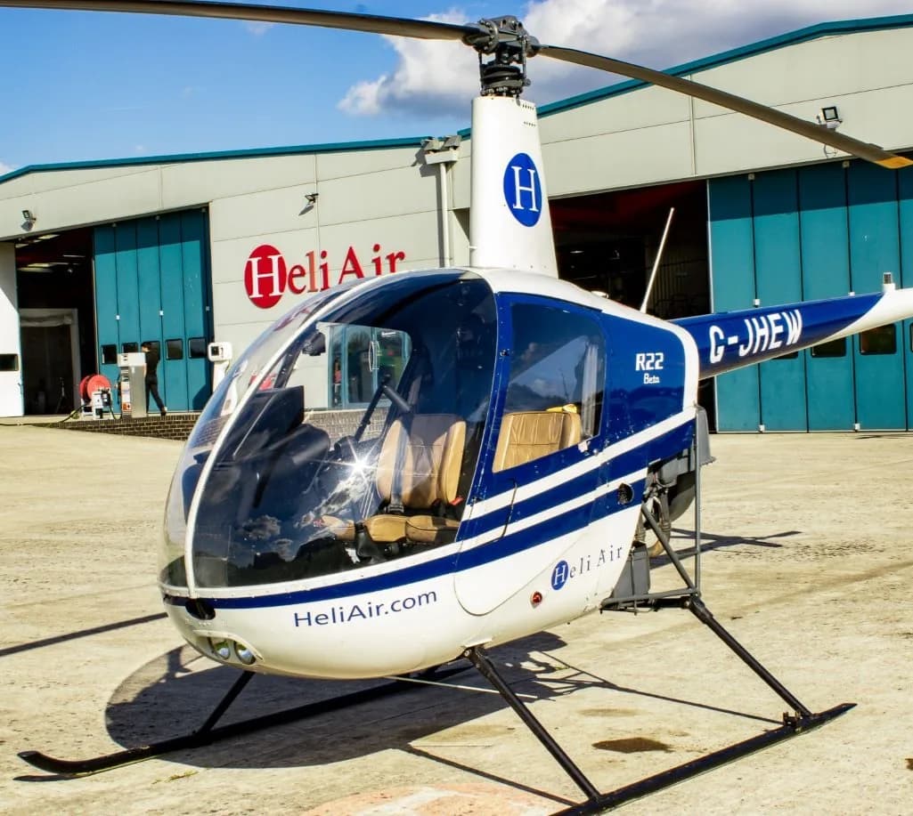A white and blue Robinson R22 Beta helicopter with "Heli Air" branding parked on an airfield in front of hangars.
