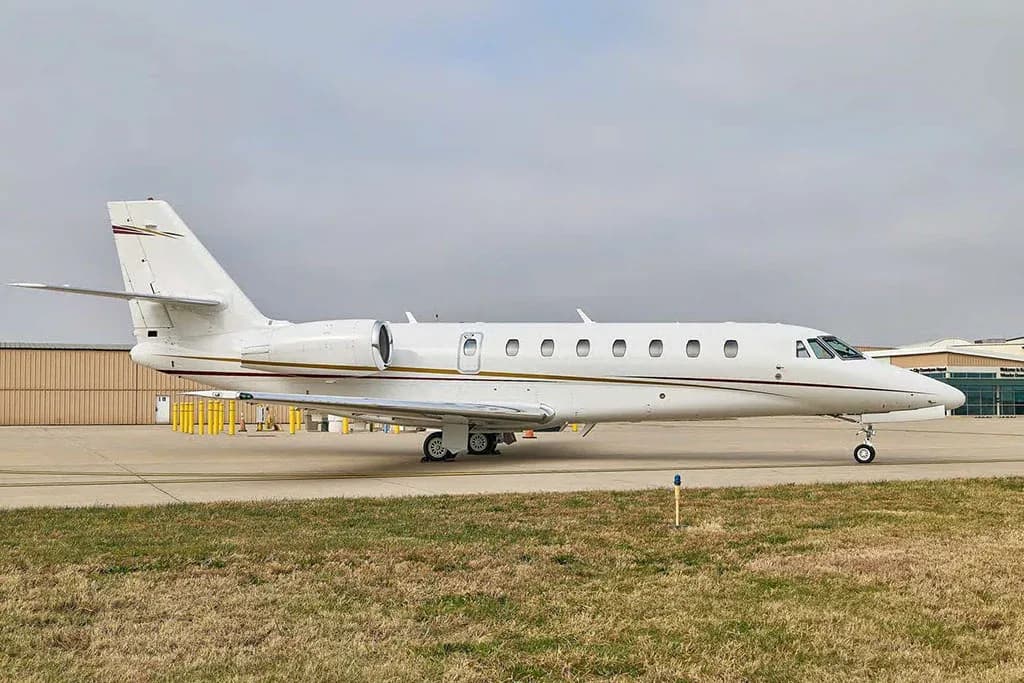 A white business jet with gold and red stripes on the tail is parked on an airport tarmac next to a building.