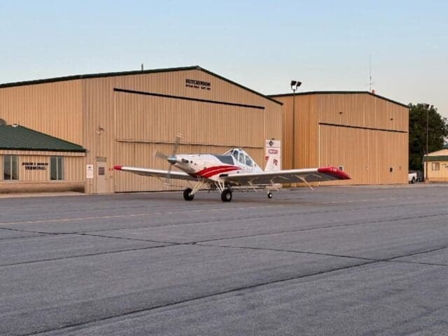A white and red agricultural aircraft is parked on an airfield ramp in front of hangars and a terminal building.